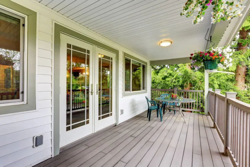 Large covered porch with railings and outdoor seats. Green trim around doors and windows of white home.