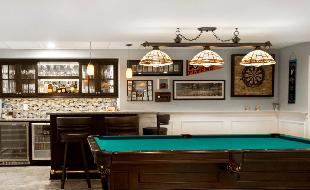 Remodeled basement with pool table, decorative pendant light above, and bar area in the background.