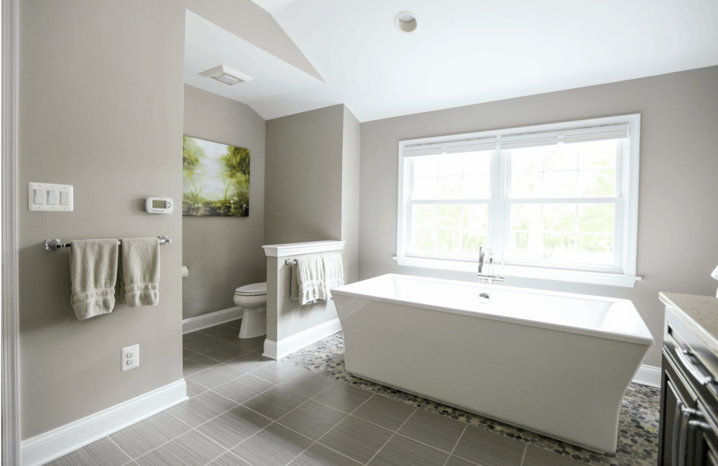 Bathroom remodel with white stand alone tub and twin double-hung windows with grids. Light gray walls and tile floor.