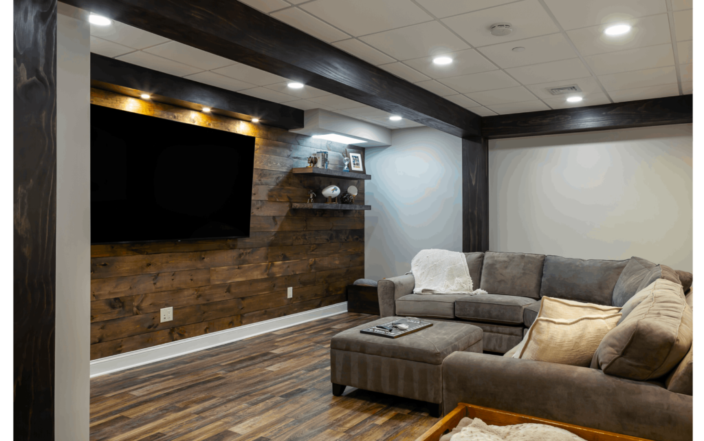 Remodeled basement with large sectional couch, wood-look floors, recessed lighting, white walls, and ship-lap wall with large TV and open shelving.