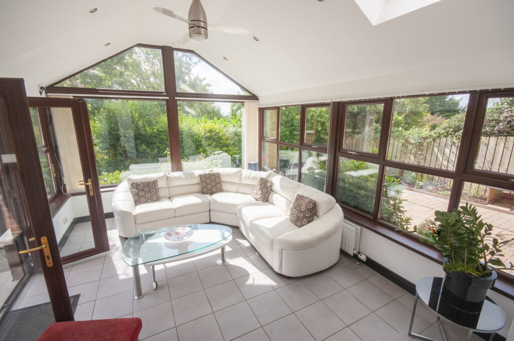 Sunroom with white tile floor, white walls with brown-trim windows, gabled ceiling, and L-shaped white couch.