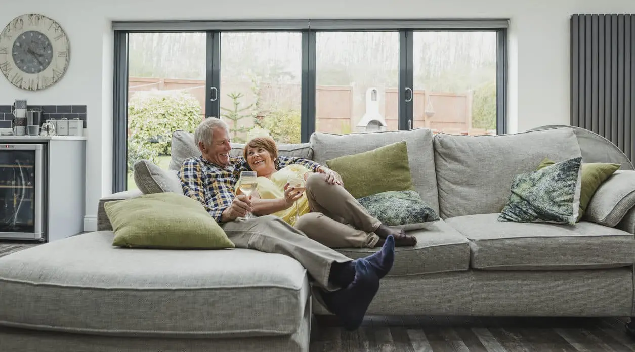 Older couple relaxing on a gray sectional sofa in a remodeled living room with double set of sliding doors in wall behind couch, leading out to a yard.