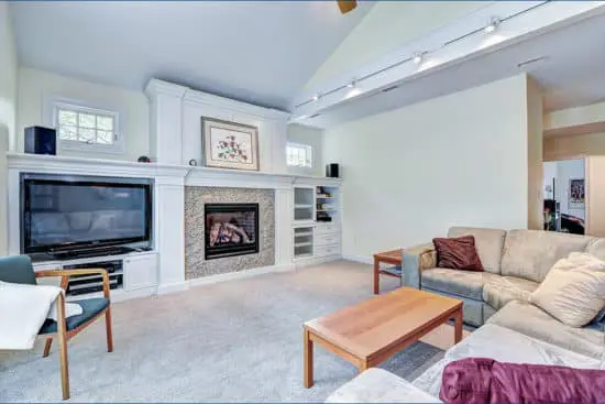 Renovated living area with track lighting and stone fireplace. White carpeted floors, white walls, white built-in shelves around fireplace.