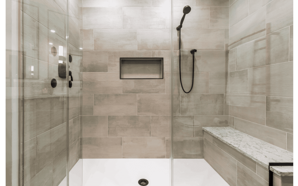 Remodeled glass-enclosed large walk-in shower with white floor tiles, large beige wall tiles, and long bench along right side. Matte black fixtures and detachable showerhead.
