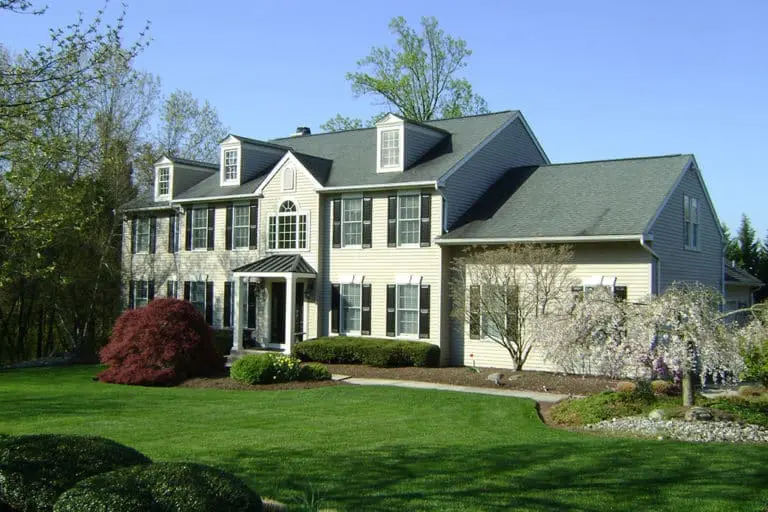 Traditional large two-story home with expansive portico and two-story addition. Three dormers. Black shutters. Landscaped front yard and trees along far side.