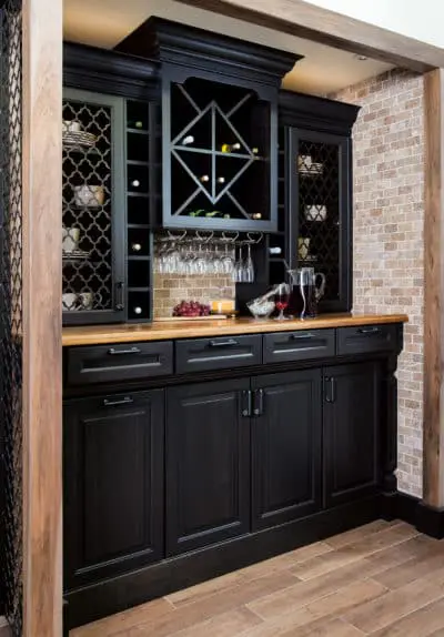 Wellborn Cabinet black cabinetry in a home bar area with light wood countertop, light brick backsplash and wall, and light wood floors.
