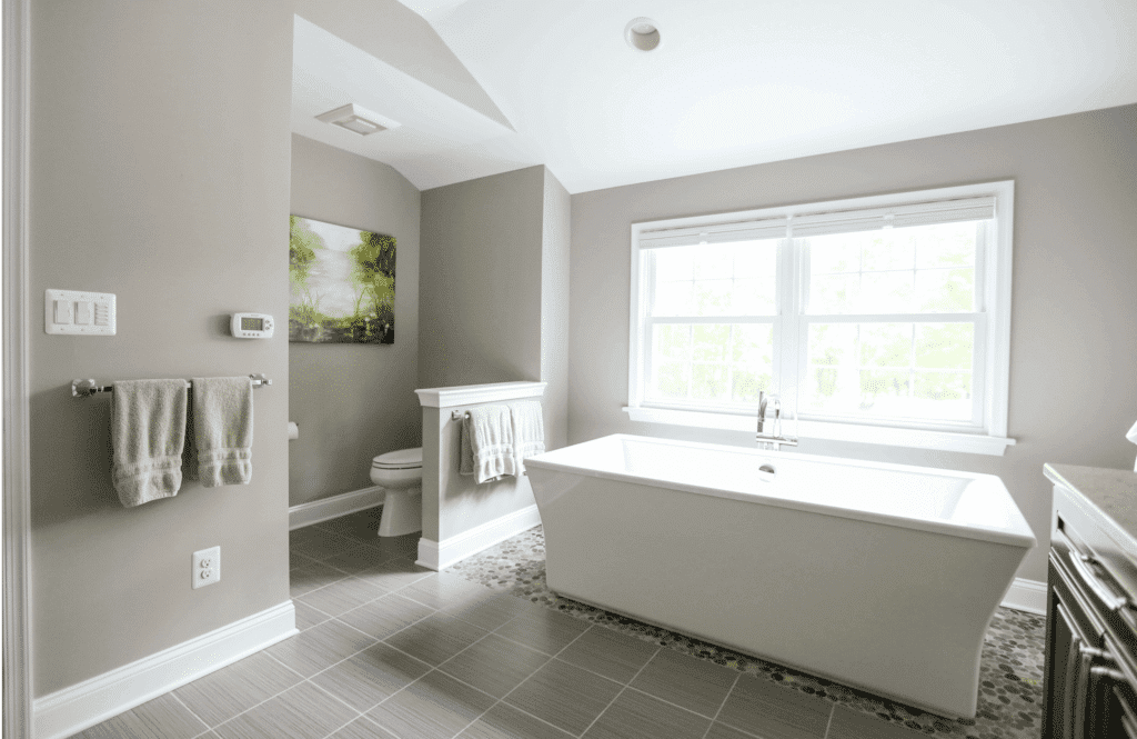 Bathroom remodel with white stand alone tub and twin double-hung windows with grids. Light gray walls and tile floor.
