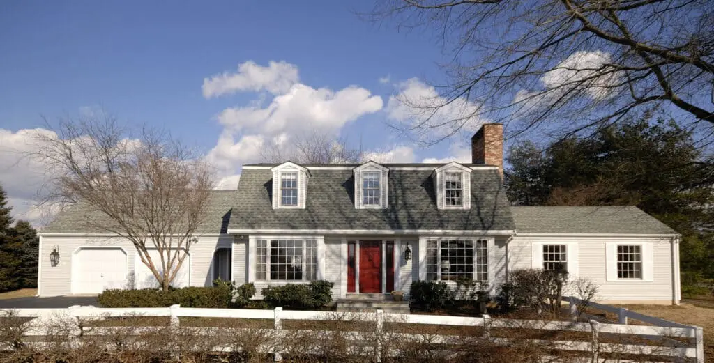 Cape Cod style home with light gray/white siding, gable roof with three dormer windows, two-car garage addition on the left. Red front door and small white fence bordering front yard.