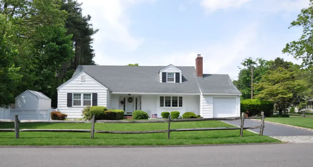 Small white ranch style home with black shutters, one small dormer window, one-car garage, and small front yard with wooden fence. Trees surrounding.
