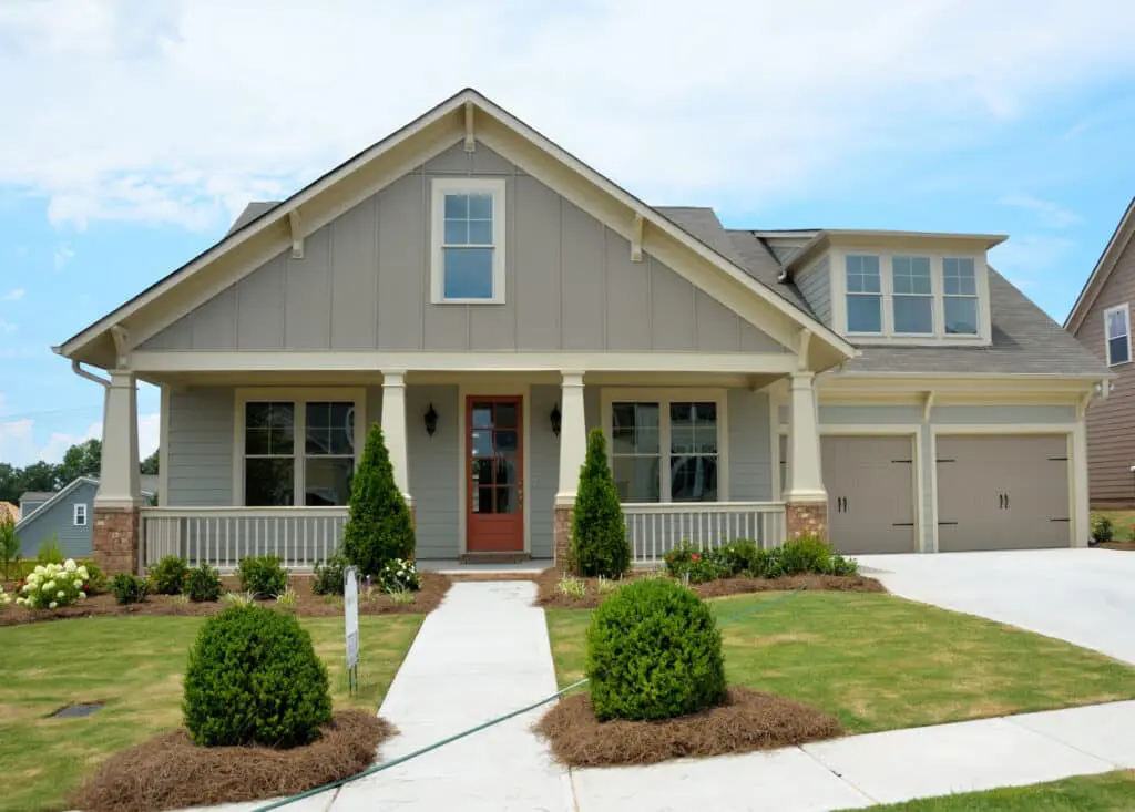 American bungalow style home exterior. Tan home with vertical siding, large front porch with wide brick-bottomed columns, two-car garage to the right. Small landscaped front yard.