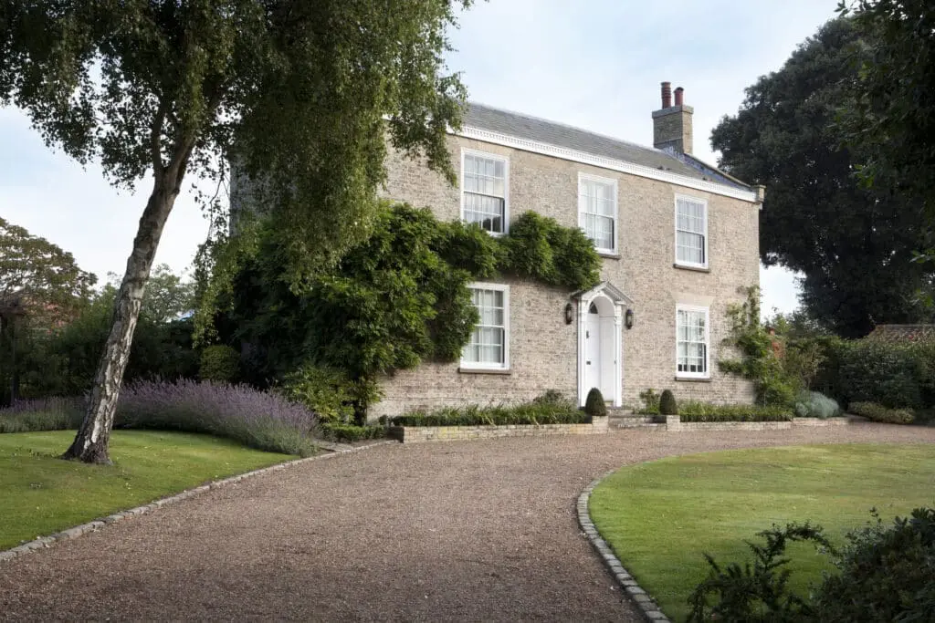 Georgian style home exterior. Large square two-story home with light tan brick and double-hung windows with grids. Curved gravel driveway in front, and trees on either side.