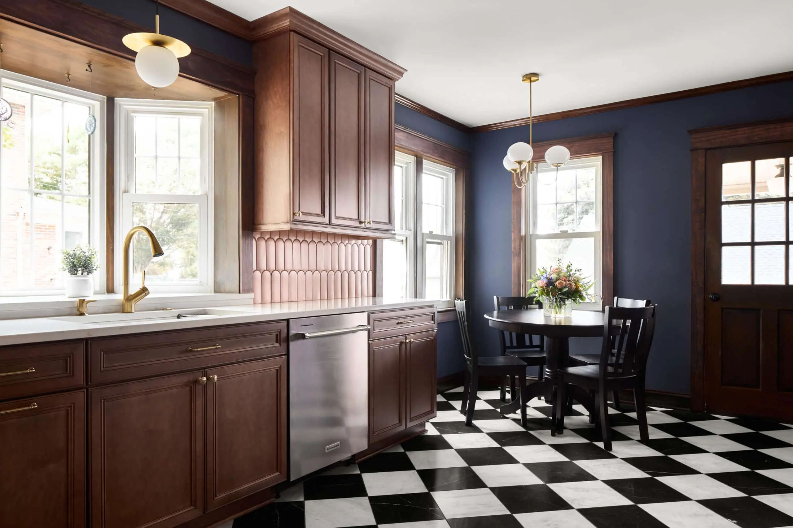 Remodeled kitchen with dark wood cabinets, white stone countertop with gold sink faucet, bay window over the sink. Black and white checked tile floor, round black dining set in far corner. Dark blue walls.
