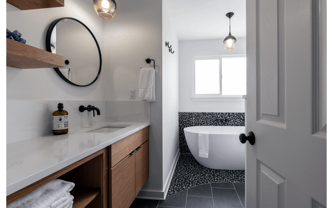 Cozy renovated bathroom with floating wooden vanity and shelves, and unique black pebble-tile under a large soaking tub