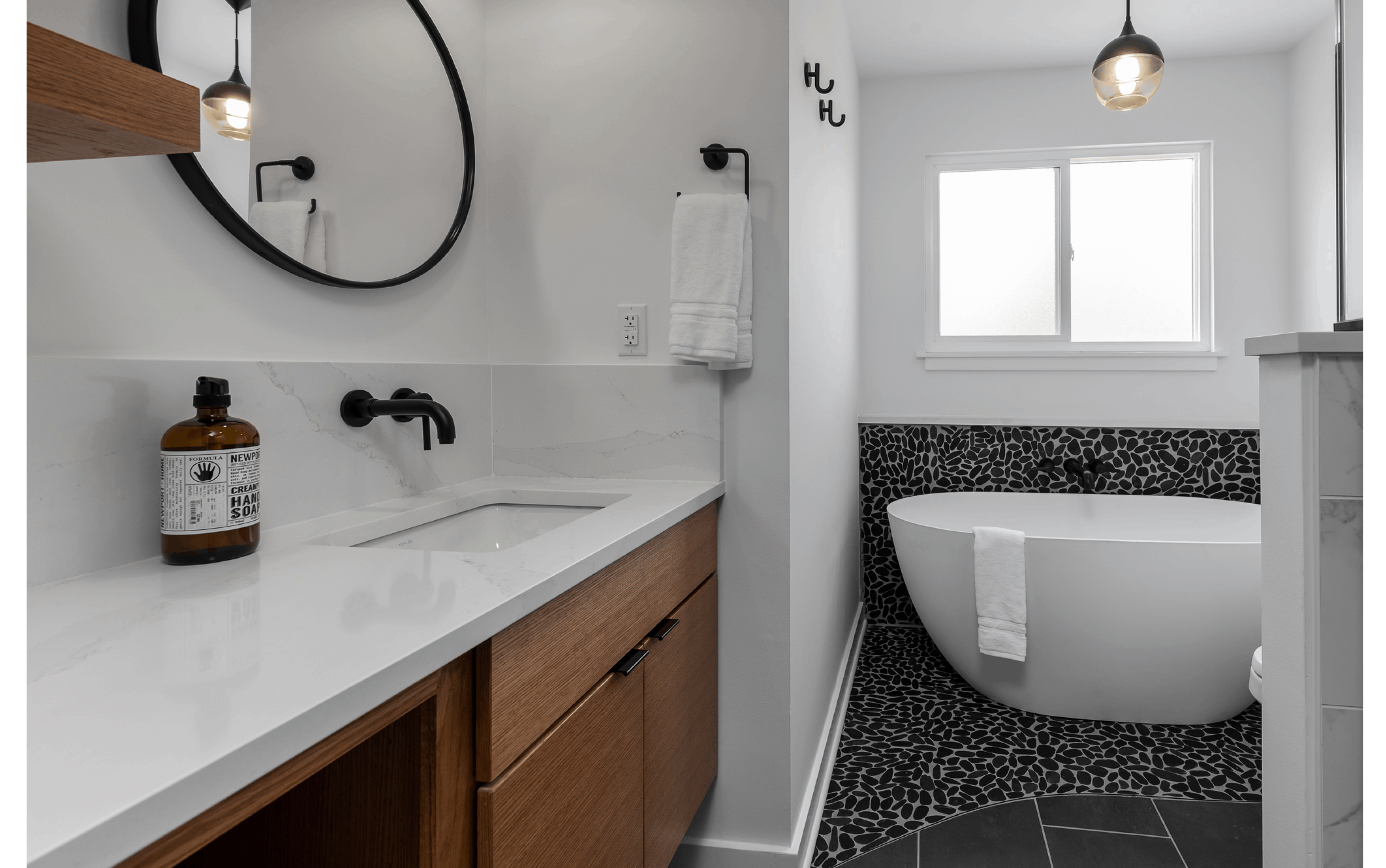 Cozy renovated bathroom with floating wooden vanity and shelves, and unique black pebble-tile under a large soaking tub