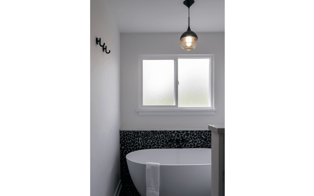 Large soaking tub sitting under rain glass window, with statement black pebble-tile on the bottom half of the wall