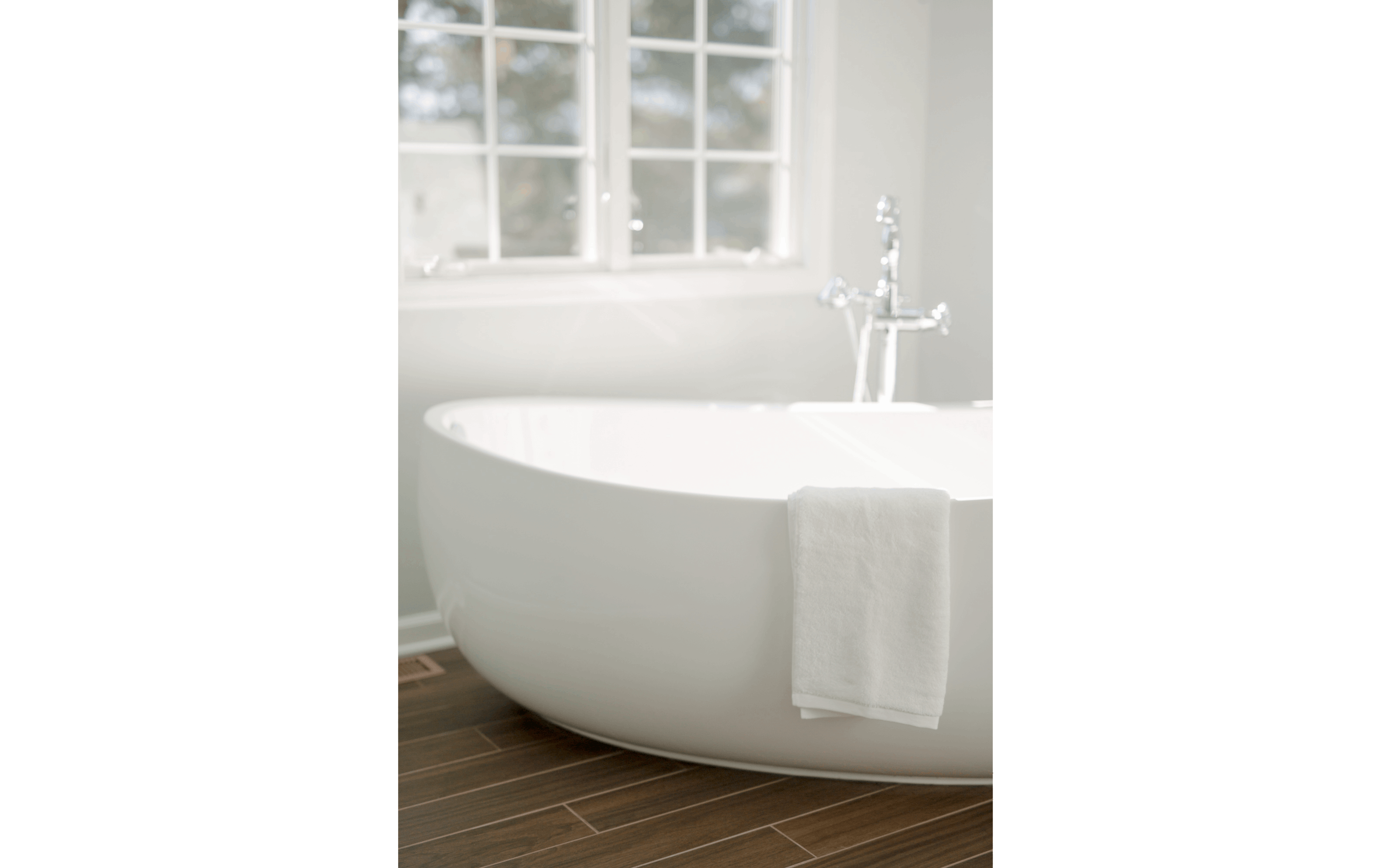 Closeup of large freestanding tub sitting against dark hardwood floors and white-trimmed double windows